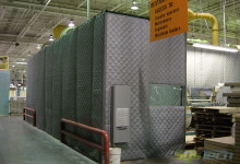 A flexible quilted blanket or soft wall noise enclosure, 55’ long x 38’ wide x 10’ tall, with floor mounted support structure, access doors, windows and a control panel ventilation point.